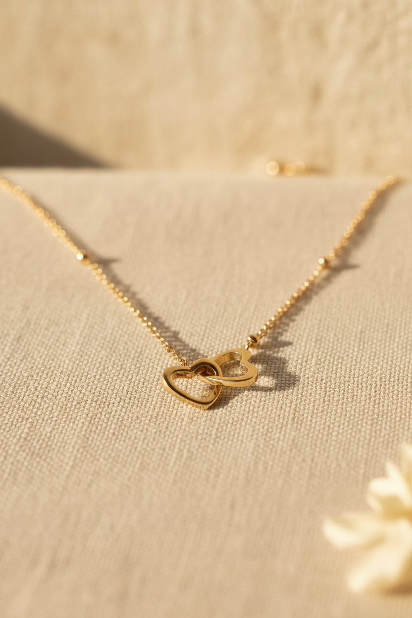Glow Mystic in Style minimalistische goudkleurige stainless steel ketting met ineengestrengelde hartjes liggend op beige stof