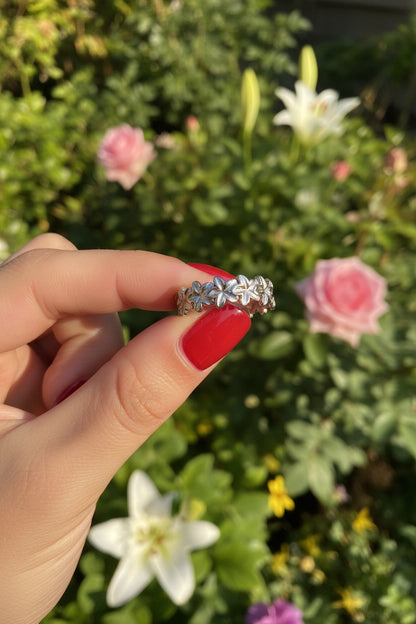 Zilverkleurige RVS smalle ring met opengewerkte bloemetjes subtiel vastgehouden tussen de vingers in de bosrijke natuur met zachte zonnestralen.