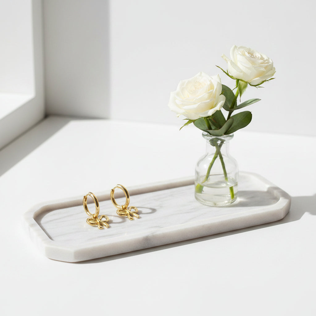 Een langwerpige marmeren tray waarop een paar gouden oorringen met een subtiel strikjes hangertje liggen. Ernaast staat een klein glazen vaasje met twee witte rozen.