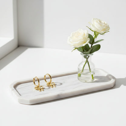Een langwerpige marmeren tray waarop een paar gouden oorringen met een subtiel strikjes hangertje liggen. Ernaast staat een klein glazen vaasje met twee witte rozen.