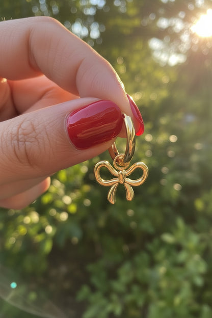 Trendy gouden waterproof RVS bow hoop oorbellen met strikje, vastgehouden in de hand in de natuur met sfeervol, warm zonlicht en een luxe esthetische uitstraling.