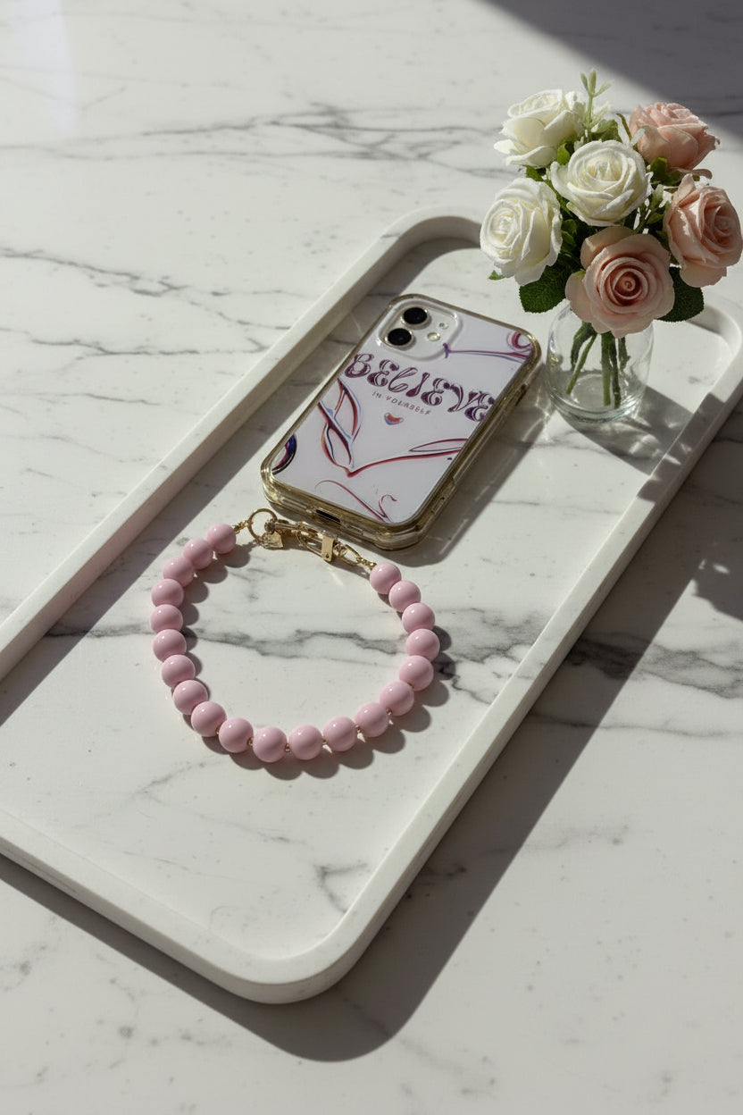 Een kort telefoonkoord van glanzende roze ronde kralen met een goudkleurige sluiting, bevestigd aan een Iphone in een transparant hoesje met de tekst 'Believe in Yourself, gepresenteerd op een langwerpig marmeren dienblad naast een glazen vaasje met witte en roze rozen.