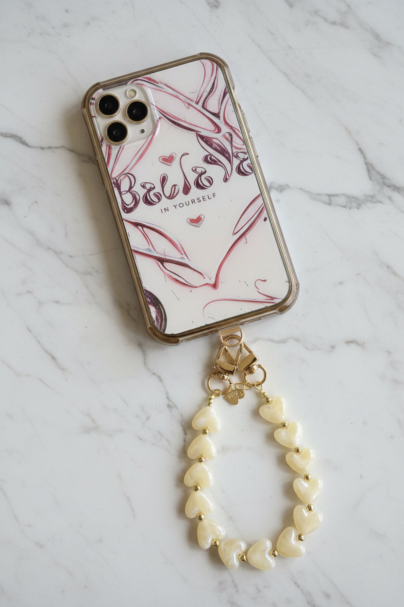 Luxe beige telefoonkoord gemaakt van glanzende parelmoer hartjeskralen en goudkleurige details met een hartjesbedel in de telefoon aangesloten op een marmeren achtergrond.