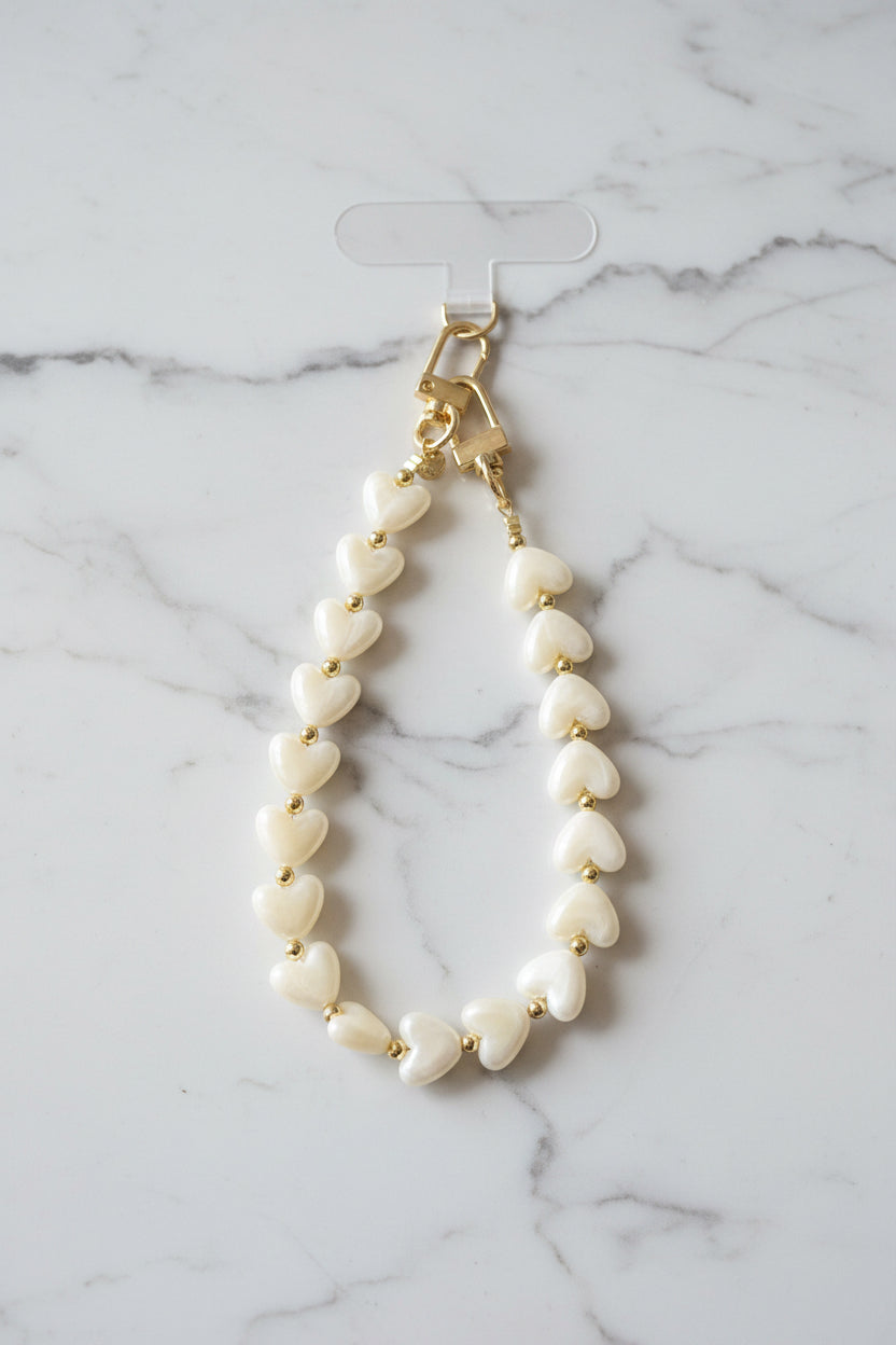 Luxe beige telefoonkoord gemaakt van glanzende parelmoer hartjeskralen en goudkleurige details met een hartjesbedel gepresenteerd op een marmeren achtergrond.