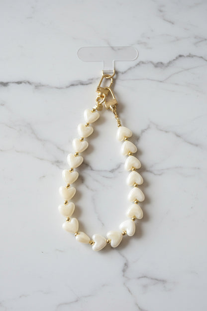 Luxe beige telefoonkoord gemaakt van glanzende parelmoer hartjeskralen en goudkleurige details met een hartjesbedel gepresenteerd op een marmeren achtergrond.