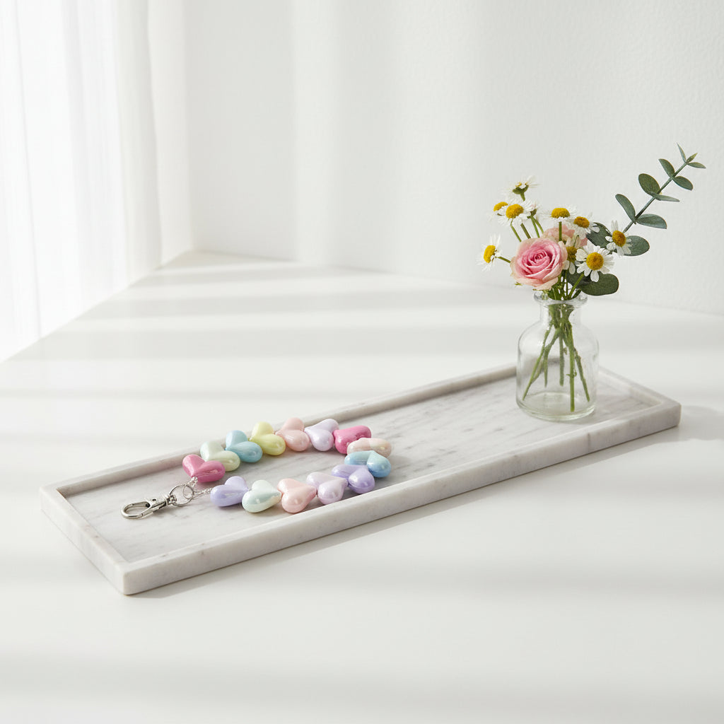 Een kort telefoonkoord van parelmoer glanzende kralen in de vorm van hartjes met een pastelkleurig verloop( roze, geel, blauw), voorzien van een zilverkleurige sluiting en gepresenteerd op een langwerpig marmeren dienblad naast een glazen vaasje met madeliefjes, een roze roos en eucalyptus.