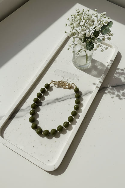 Olive green telefoonkoord met ronde kralen en goudkleurige sluiting, gepresenteerd op een marmeren dienblad naast een glazen vaasje met witte bloemen.