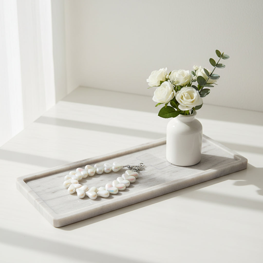 Een kort telefoonkoord gemaakt van glanzende, witte parelmoer kralen in de vorm van hartjes, voorzien van een zilverkleurige sluiting en gepresenteerd op een langwerpig wit marmeren dienblad naast een klein wit vaasje met witte rozen.