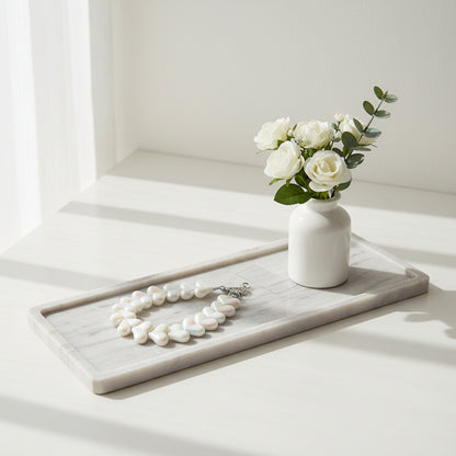 Een kort telefoonkoord gemaakt van glanzende, witte parelmoer kralen in de vorm van hartjes, voorzien van een zilverkleurige sluiting en gepresenteerd op een langwerpig wit marmeren dienblad naast een klein wit vaasje met witte rozen.