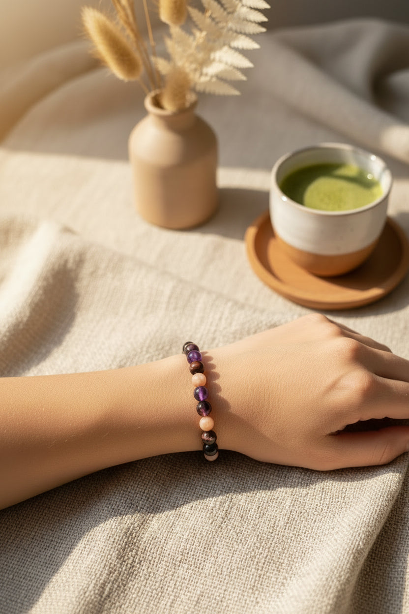 Sfeerbeeld van een hand die rust op een beige linnen doek, met een verfijnde edelsteen armband van paarse amethist, bruine tijgeroog en zachtroze zonnesteen, naast een kopje matcha thee.