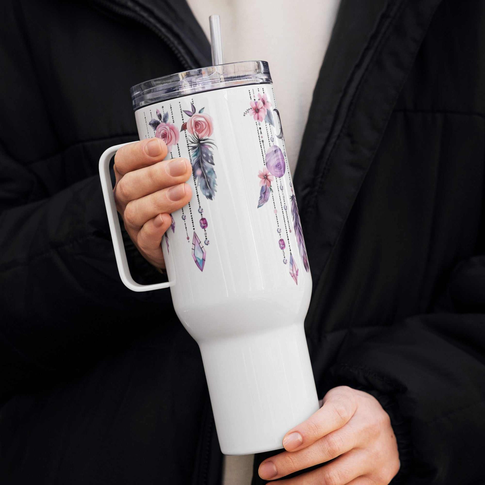 White travel mug with boho feather and floral art, insulated with handle and clear lid with straw, held by person in black jacket