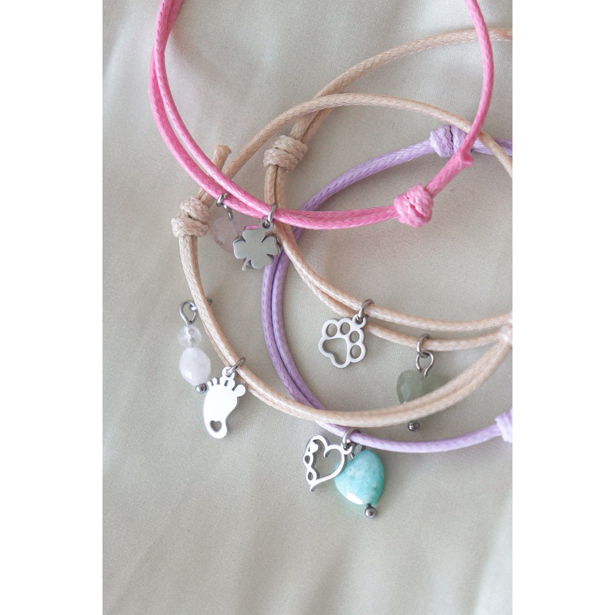 "Sfeerfoto van diverse kleurrijke waxkoord armbandjes met zilverkleurige bedeltjes zoals babyvoetjes en edelstenen, liggend op een zachte ondergrond."