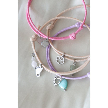 "Sfeerfoto van diverse kleurrijke waxkoord armbandjes met zilverkleurige bedeltjes zoals babyvoetjes en edelstenen, liggend op een zachte ondergrond."