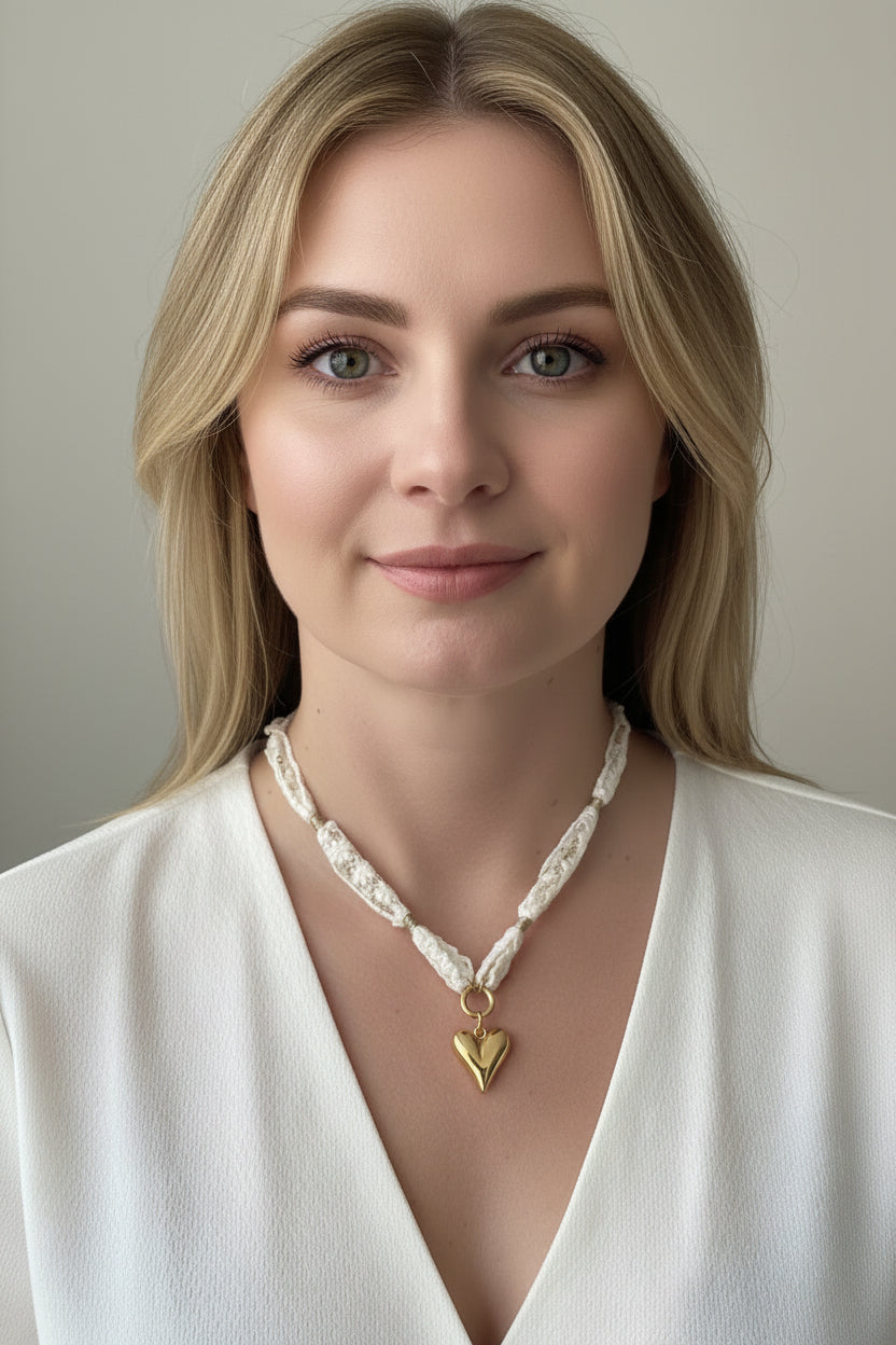 Close-up shot van de witte kanten choker met goud hart hanger gedragen op een hals van een model die een witte blouse draagt in een cleane setting.