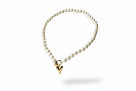 Parel-look kralenketting van Glow Mystic, plat neergelegd op een lichte ondergrond, met glanzende witte kralen en een goudkleurige Stainless Steel harthanger uit de collection."