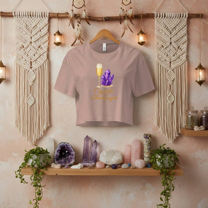 Crystals and Champagne Women's Crop T-Shirt with champagne glass and purple crystals design hanging in boho room with crystals and plants on wooden shelf