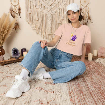 Woman wearing Crystals and Champagne Women's Crop T-Shirt with purple crystals and champagne glass design, paired with jeans and white sneakers, sitting in boho room setting