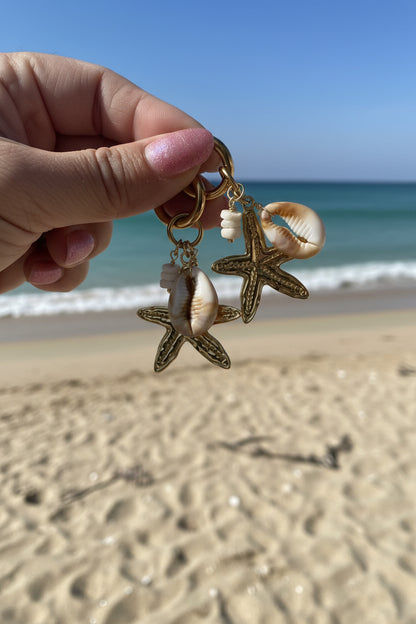 Luxe goudkleurige strandsieraden met zeester en kauri-schelp bedels tegen een achtergrond van een blauwe zee en zandstrand.