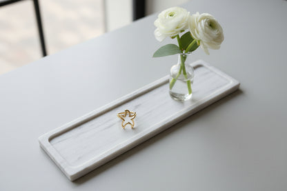 Goudkleurige opengewerkte ring in de vorm van een zeester, gepresenteerd op een marmeren plateau naast een vaasje met witte bloemen.