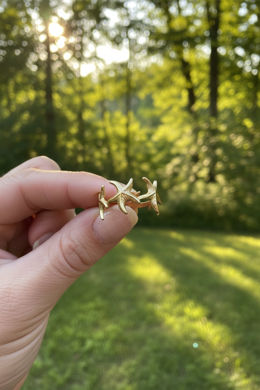 Goudkleurige verstelbare RVS zeester ring subtiel vastgehouden tussen de vingers in de bosrijke natuur met sfeervolle, glanzende zonnestralen.