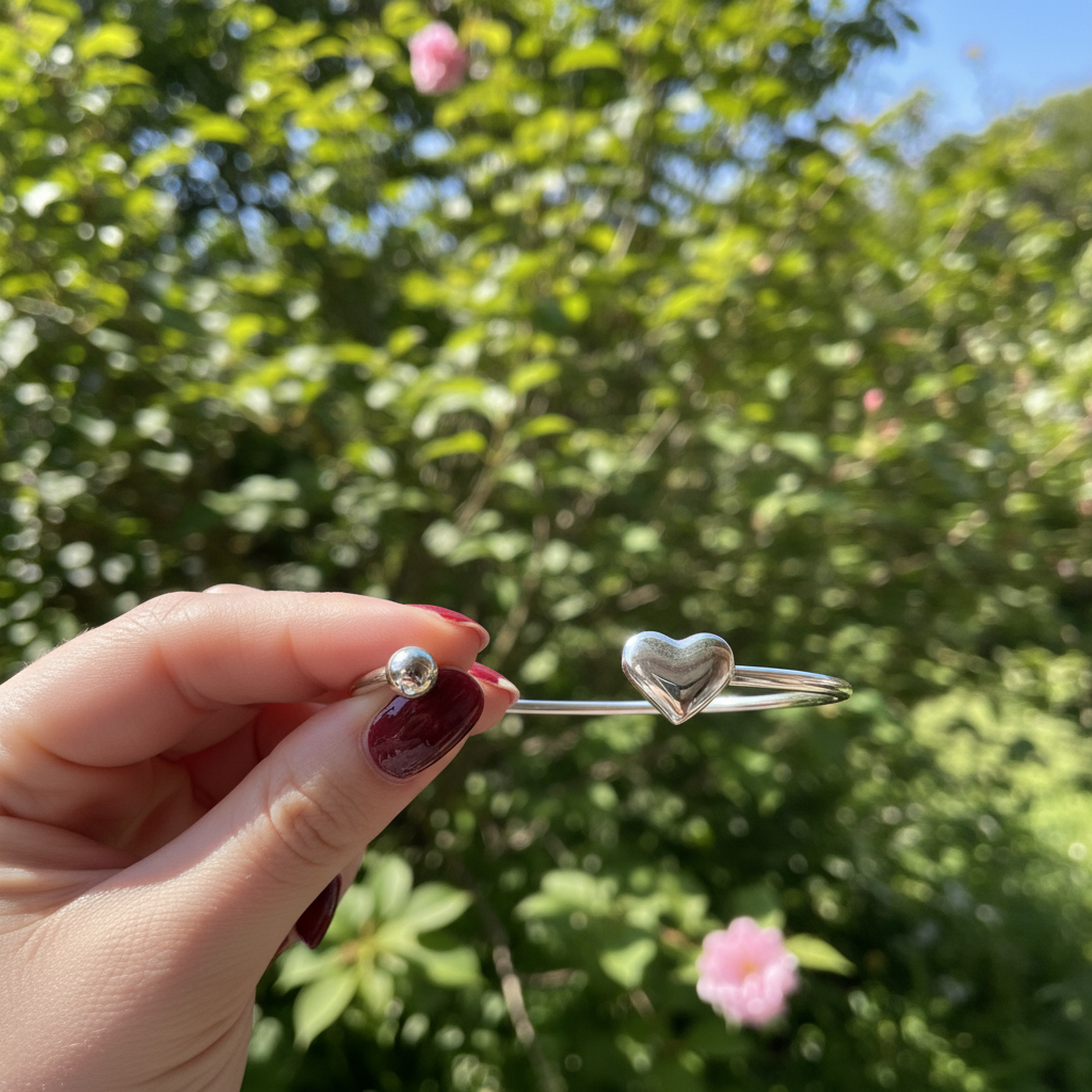 Detailopname van een minimalistische zilveren RVS-bangle met hartje, vastgehouden door een hand met donkerrode, perfect verzorgde nagels buiten in de natuur met zonlicht.