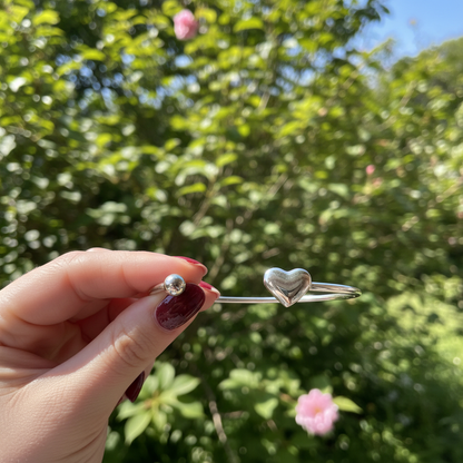 Detailopname van een minimalistische zilveren RVS-bangle met hartje, vastgehouden door een hand met donkerrode, perfect verzorgde nagels buiten in de natuur met zonlicht.